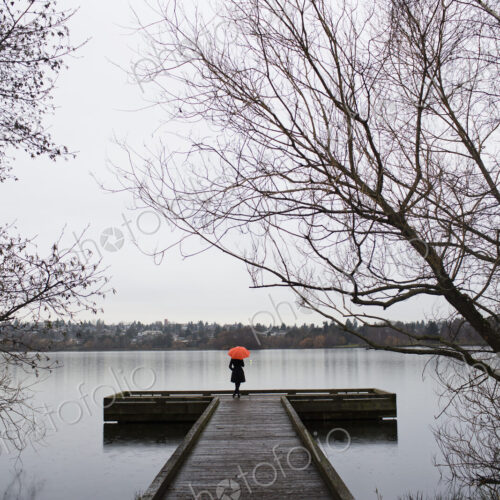 A woman standing at the end of a dock with an orange umbrella on a cloudy, grey day in…