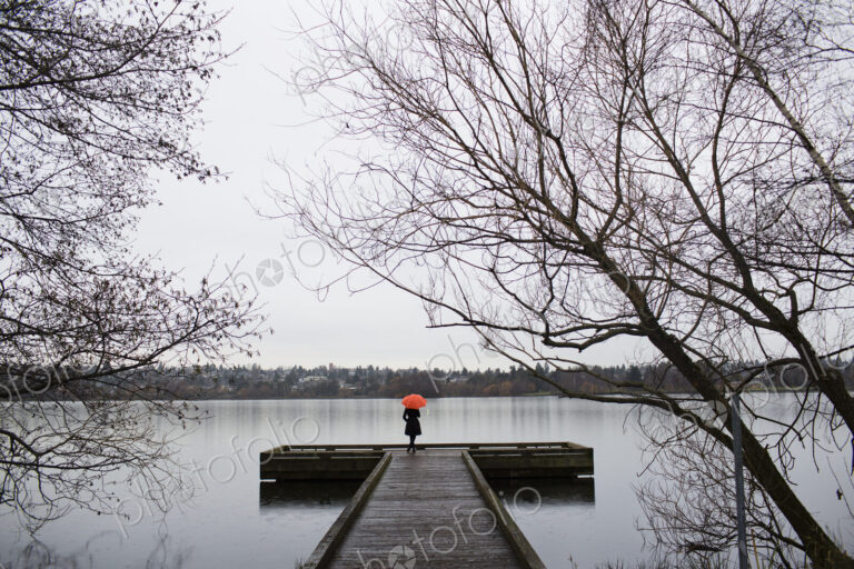 A woman standing at the end of a dock with an orange umbrella on a cloudy, grey day in…