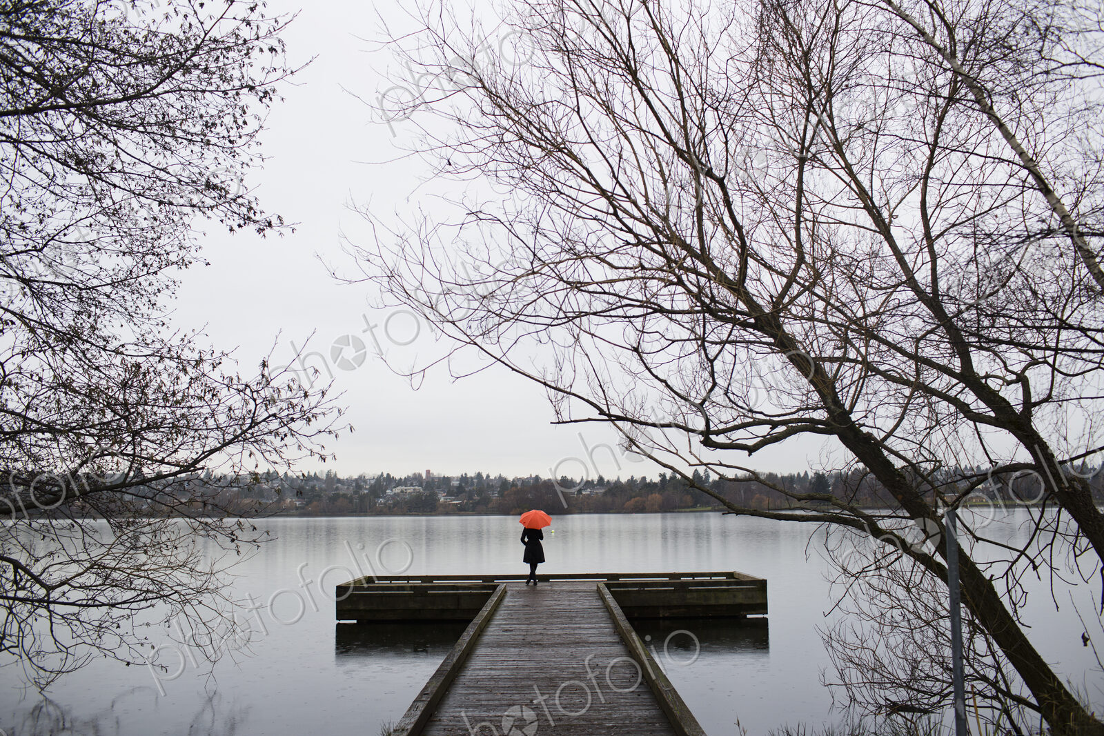 A woman standing at the end of a dock with an orange umbrella on a cloudy, grey day in…