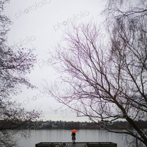 A woman standing at the end of a dock with an orange umbrella on a cloudy, grey day in…