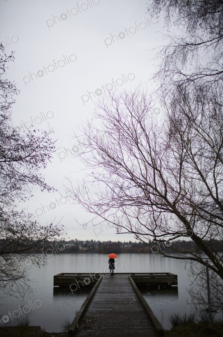 A woman standing at the end of a dock with an orange umbrella on a cloudy, grey day in…