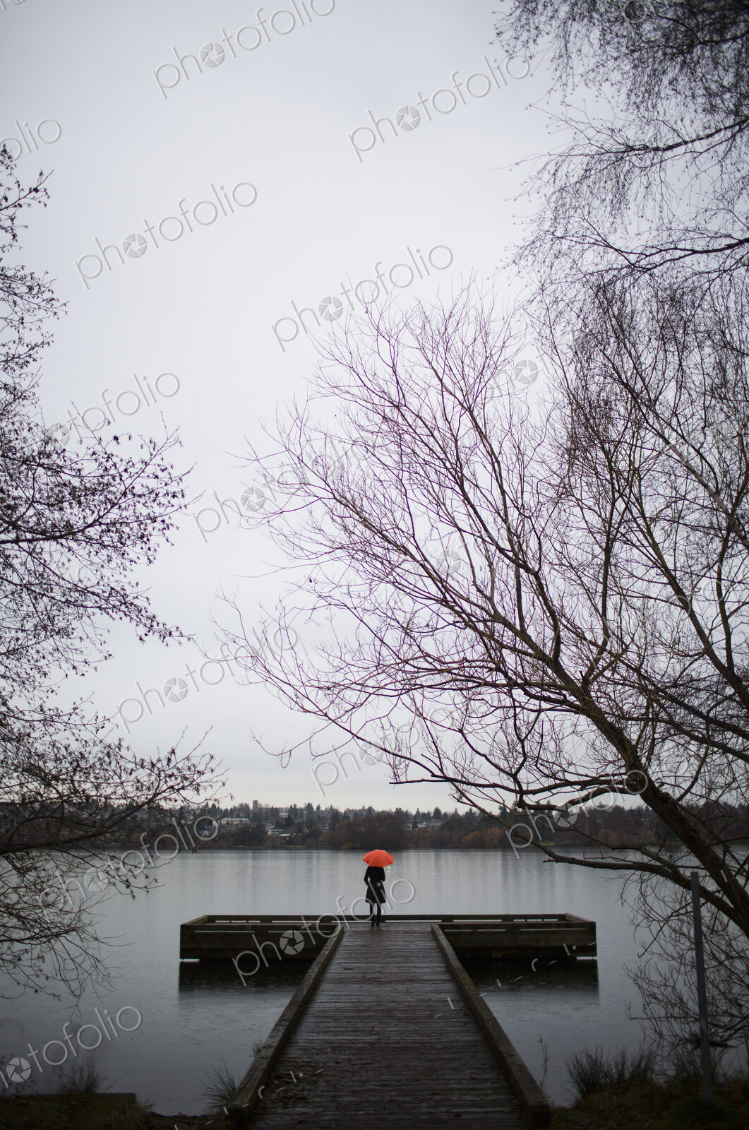 A woman standing at the end of a dock with an orange umbrella on a cloudy, grey day in…