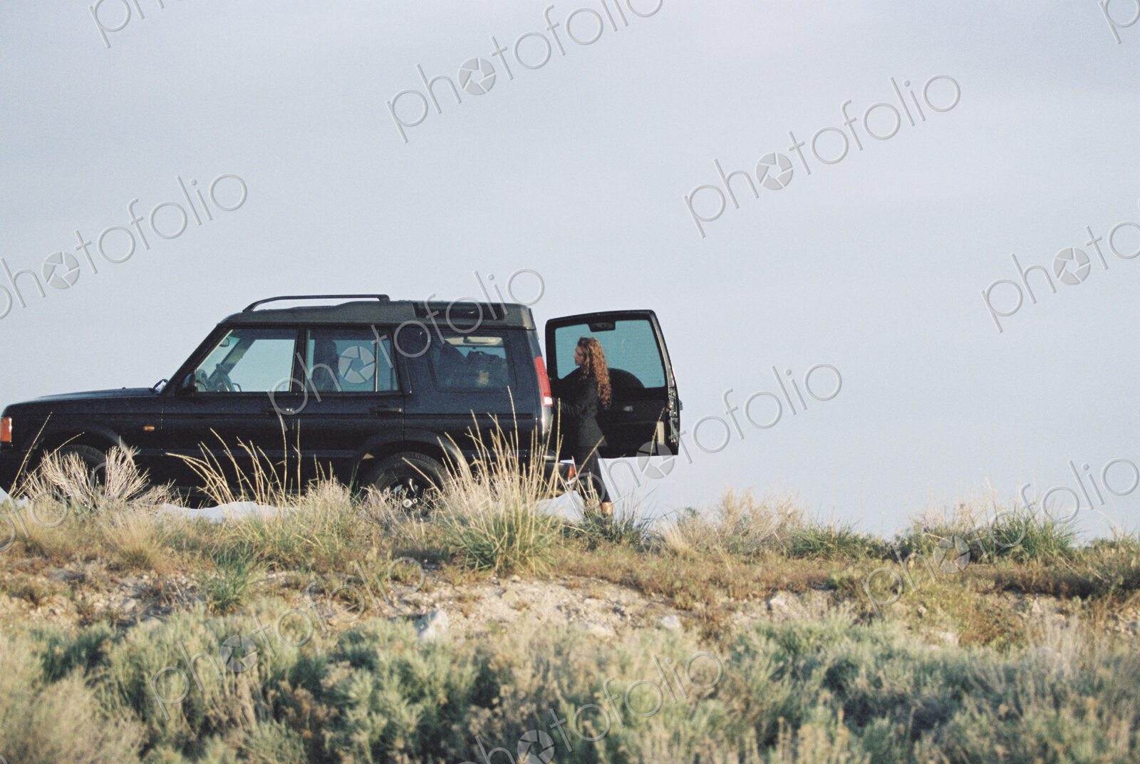 A woman standing by the open rear door of a 4×4 in open space.