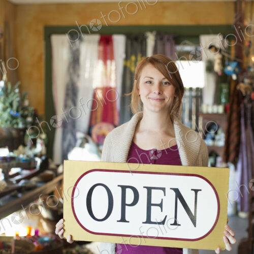 A woman standing in an antique store, holding an OPEN sign. Displays of goods all… A woman standing in an antique store, holding an OPEN sign. Displays of goods all…