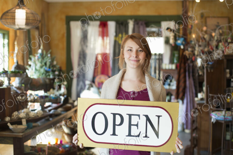 A woman standing in an antique store, holding an OPEN sign. Displays of goods all…