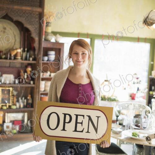 A woman standing in an antique store, holding an OPEN sign. Displays of goods all… A woman standing in an antique store, holding an OPEN sign. Displays of goods all…