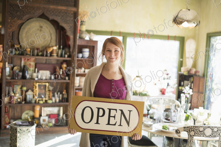 A woman standing in an antique store, holding an OPEN sign. Displays of goods all…