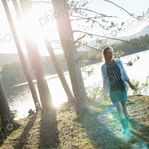 A woman standing in the shadows of tall trees on the shores of a lake in summer. A woman standing in the shadows of tall trees on the shores of a lake in summer.