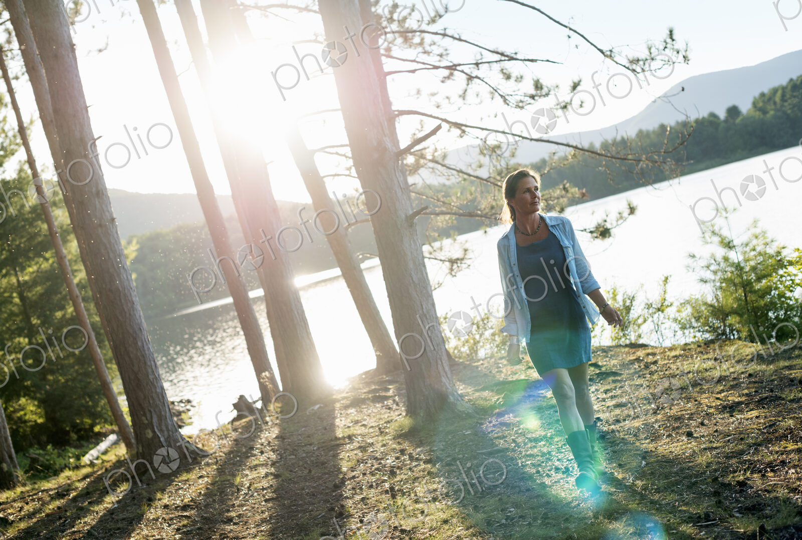A woman standing in the shadows of tall trees on the shores of a lake in summer.