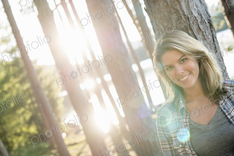 A woman standing in the shadows of tall trees on the shores of a lake in summer.
