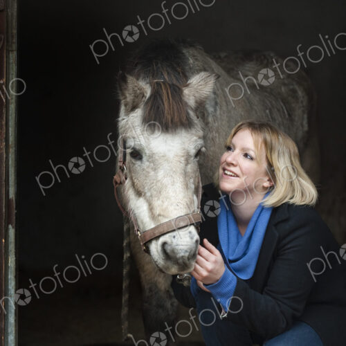 A woman standing next to a grey horse stroking its muzzle, at the stable door. A woman standing next to a grey horse stroking its muzzle, at the stable door.