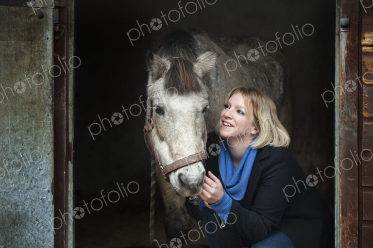 A woman standing next to a grey horse stroking its muzzle, at the stable door.