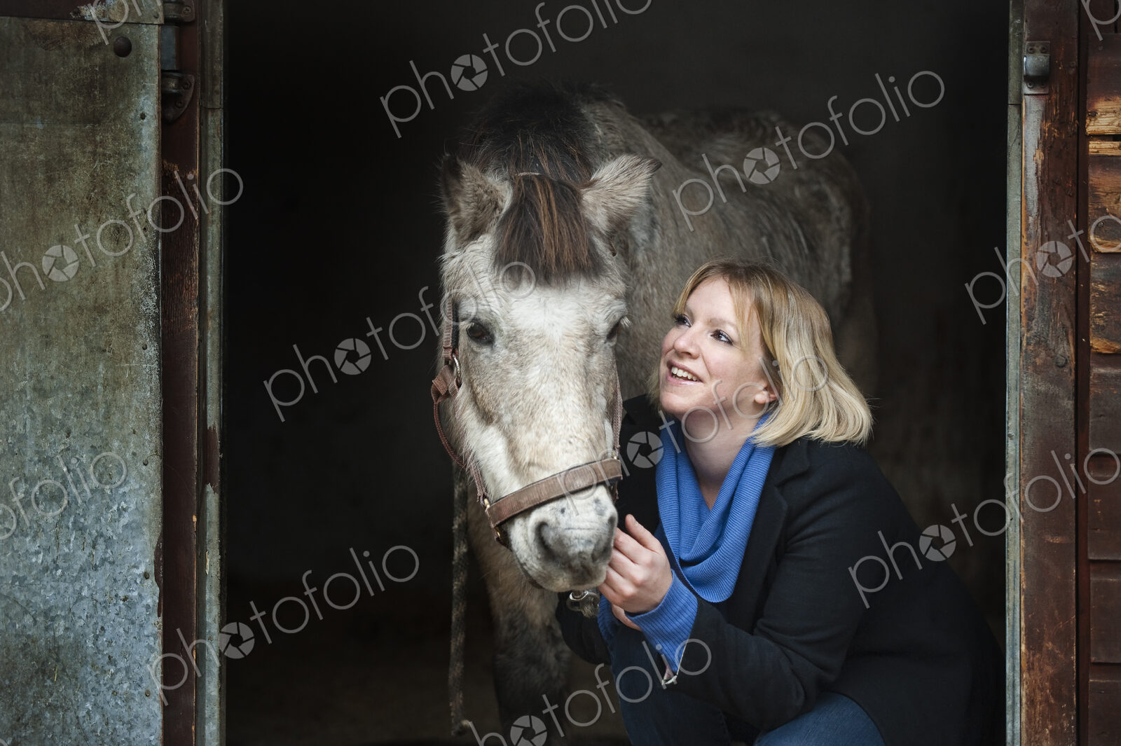A woman standing next to a grey horse stroking its muzzle, at the stable door.