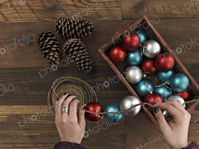 A woman threading Christmas shiny round ornaments on a piece of string. A small group…