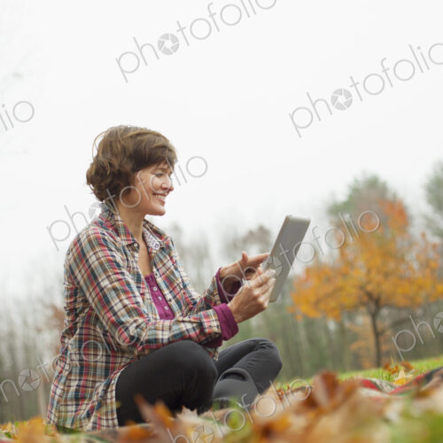 A woman using a computer tablet sitting on the ground. Autumn leaves and woodland.