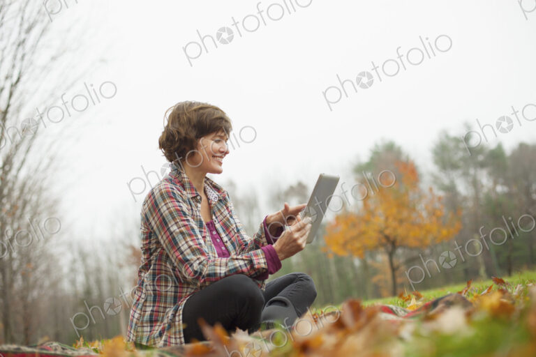 A woman using a computer tablet sitting on the ground. Autumn leaves and woodland.