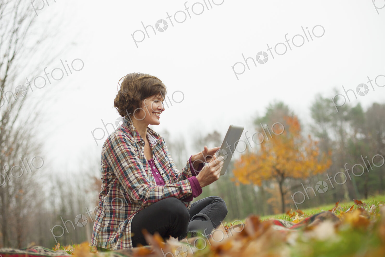 A woman using a computer tablet sitting on the ground. Autumn leaves and woodland.