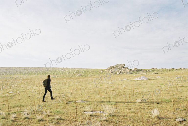 A woman walking across open ground towards a small cairn, carrying a backpack.