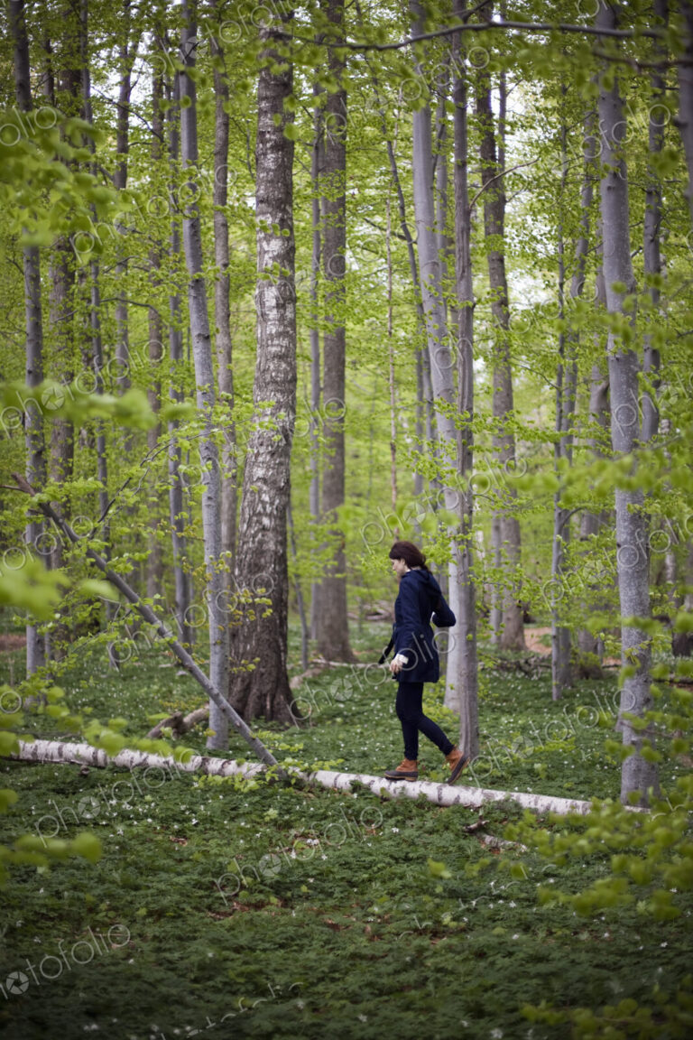A woman walking along a fallen tree trunk in the woods. Balancing on the narrow piece…