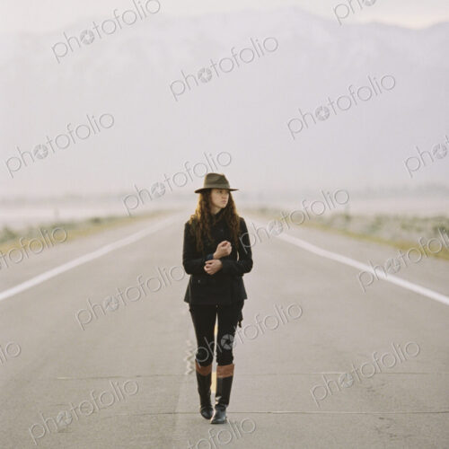A woman walking down the middle of a country road, wearing a hat and carrying a backpack.