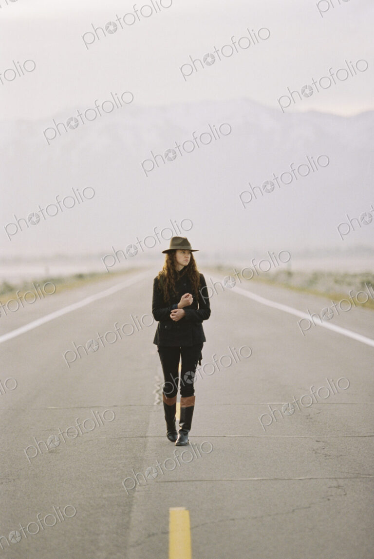 A woman walking down the middle of a country road, wearing a hat and carrying a backpack.