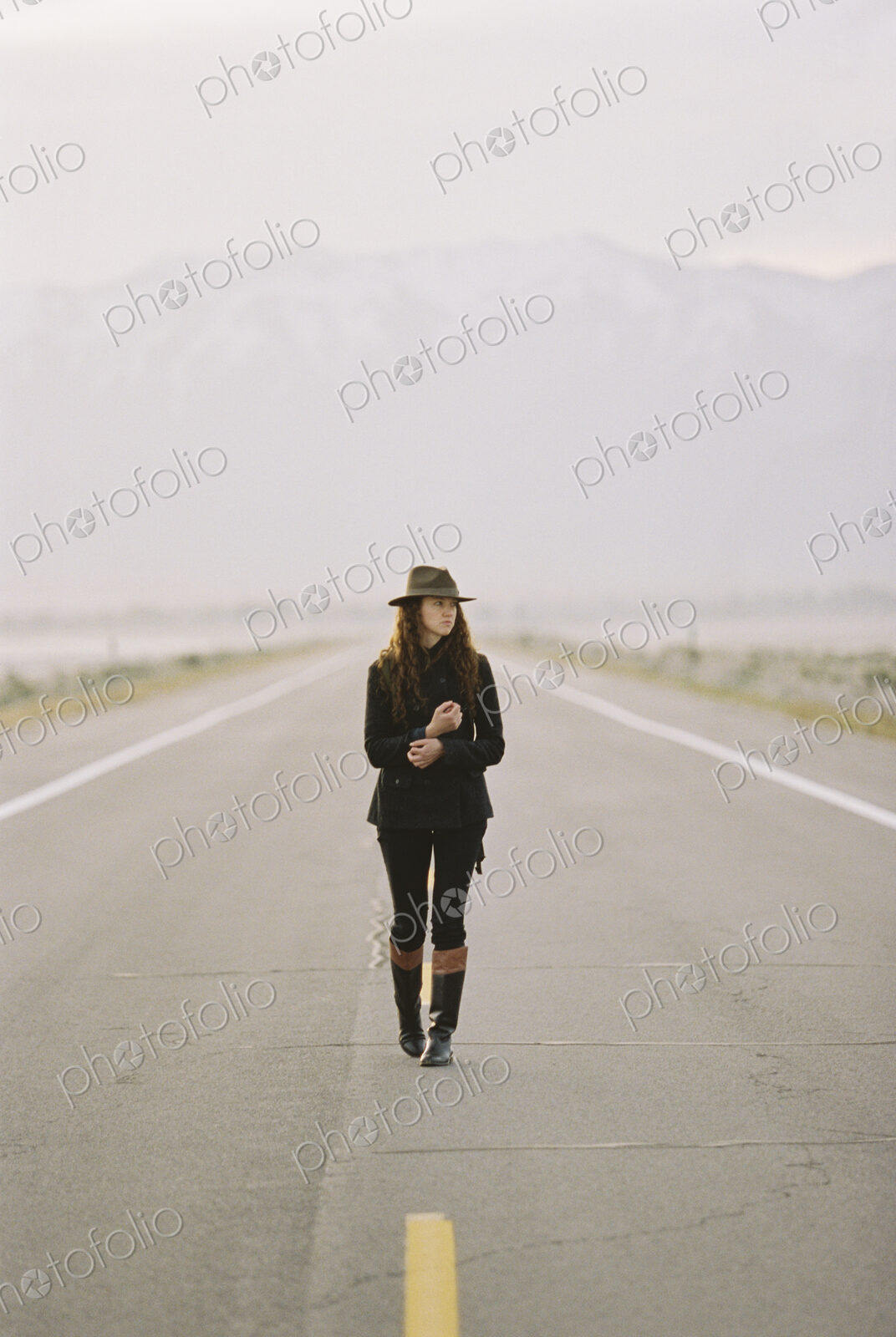 A woman walking down the middle of a country road, wearing a hat and carrying a backpack.