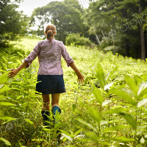 A woman walking through the undergrowth in woodland, with her arms brushing the tops…