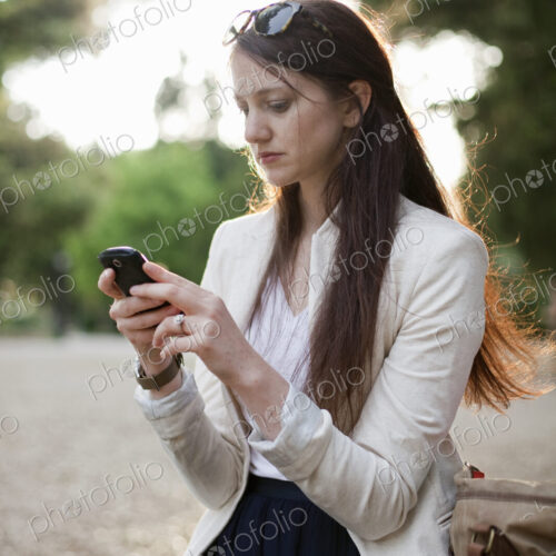 A woman wearing a cream jacket, sitting in a city park looking at her cell phone.