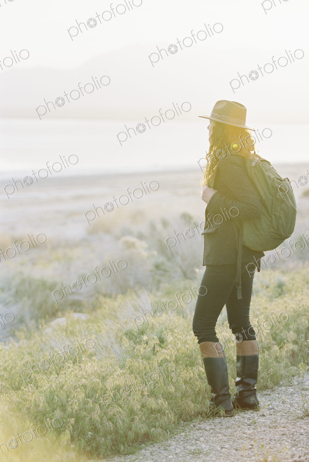 A woman wearing a hat and carrying a backpack, standing on a path overlooking a lake…