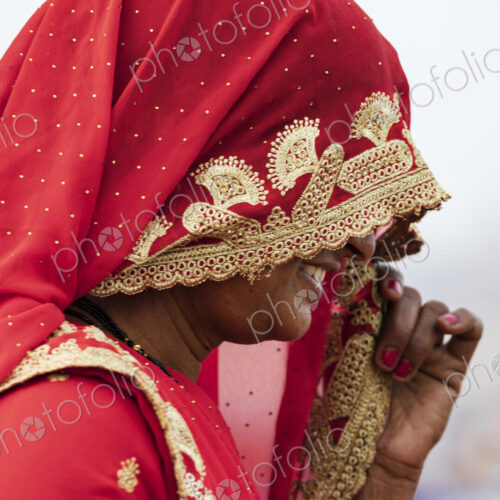 A woman wearing a red sari and head covering decorated with golden thread and flower…