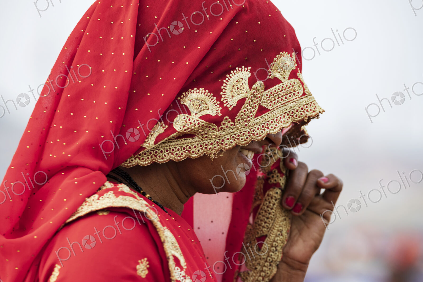 A woman wearing a red sari and head covering decorated with golden thread and flower…
