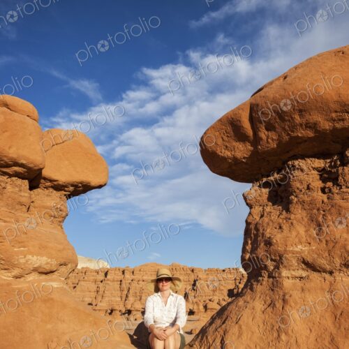 A woman wearing a wide-brimmed hat and sunglasses sits barefoot on warm red sandstone…