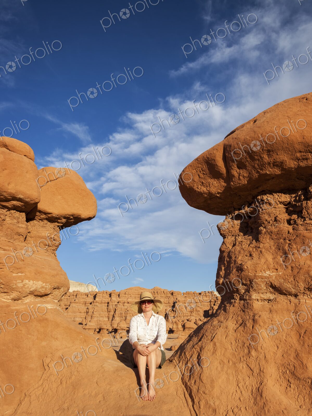 A woman wearing a wide-brimmed hat and sunglasses sits barefoot on warm red sandstone…
