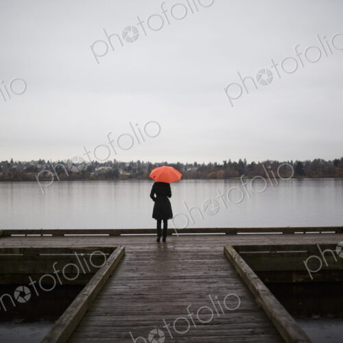 A woman wearing all black and holding an orange umbrella stands on the edge of a dock…