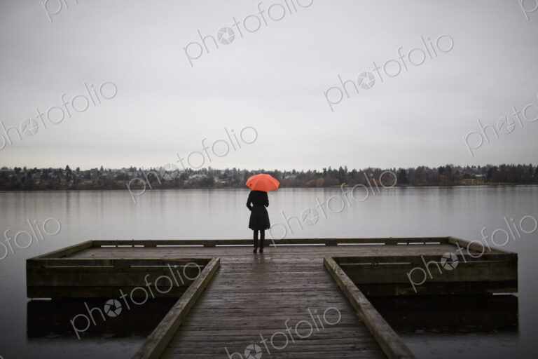 A woman wearing all black and holding an orange umbrella stands on the edge of a dock…
