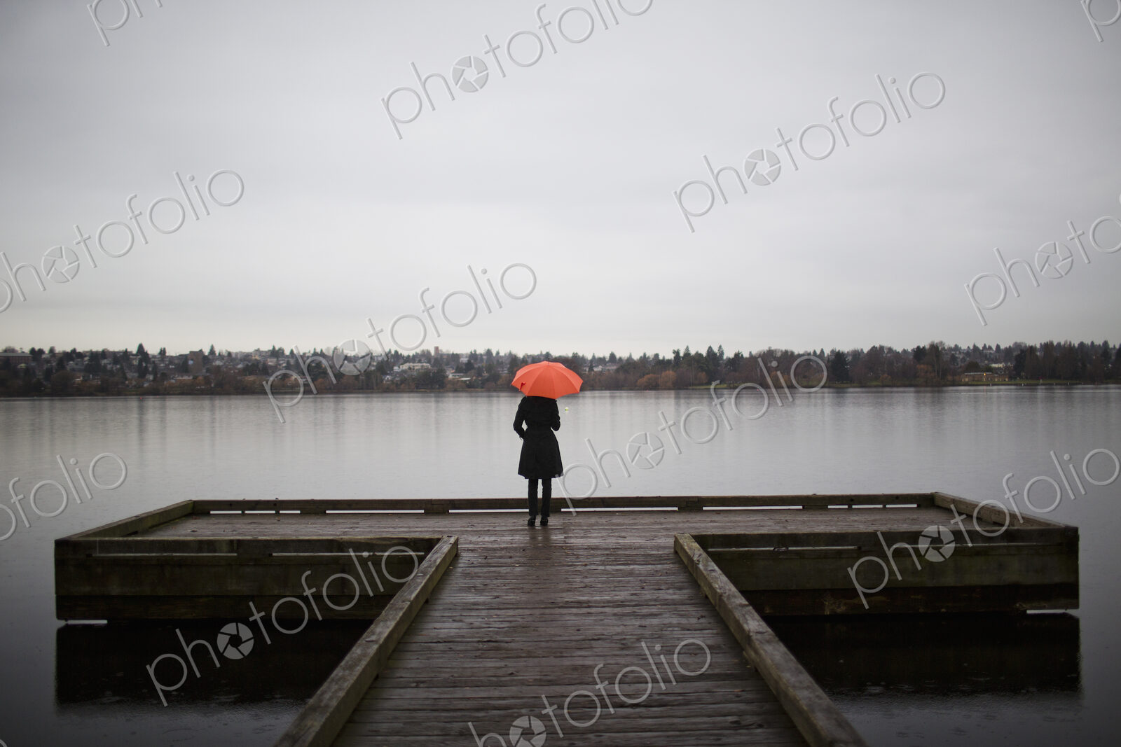 A woman wearing all black and holding an orange umbrella stands on the edge of a dock…