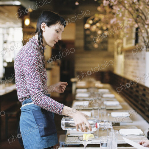 A woman wearing an apron pouring water into glasses on a long narrow table A woman wearing an apron pouring water into glasses on a long narrow table