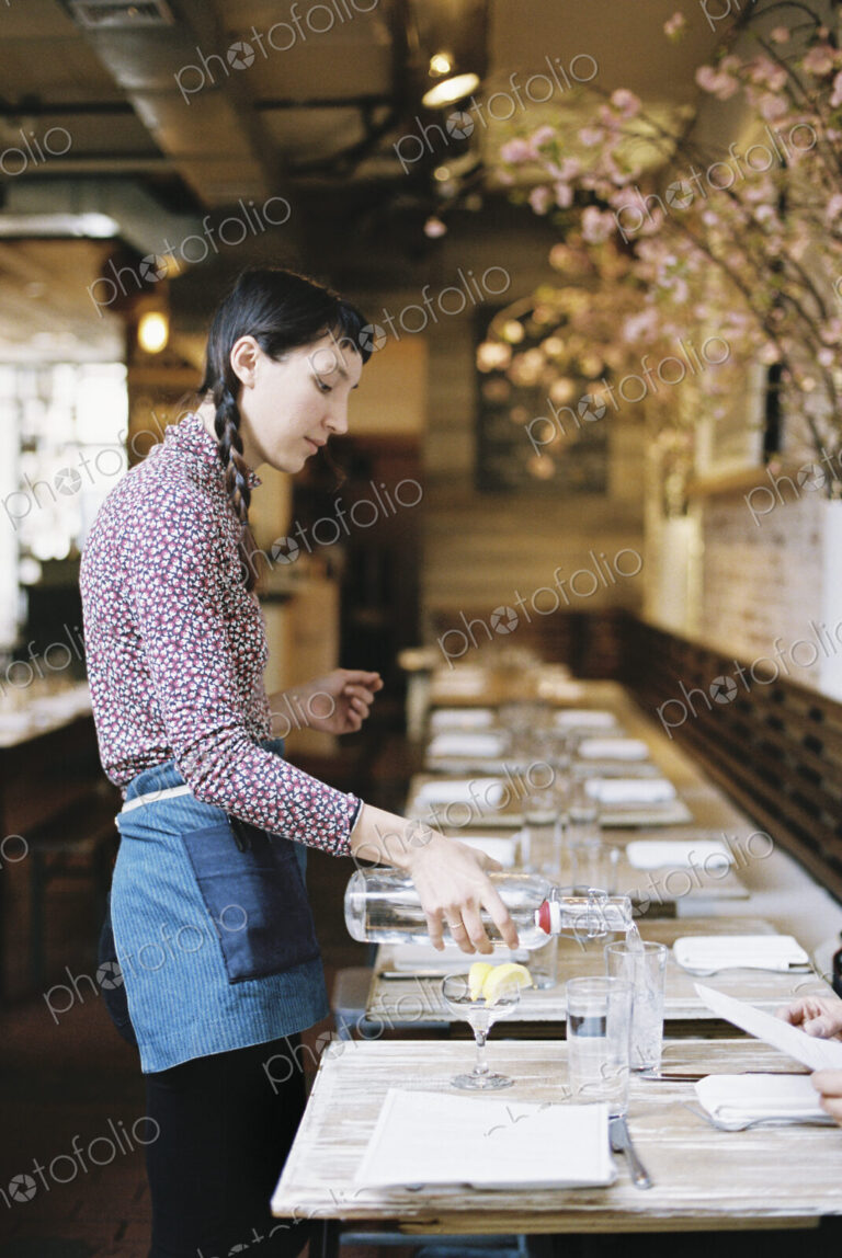 A woman wearing an apron pouring water into glasses on a long narrow table