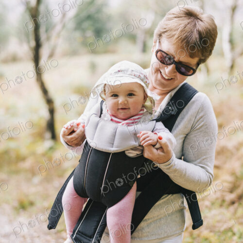 A woman wearing sunglasses carrying a baby wearing a white sunhat in a front baby sling.