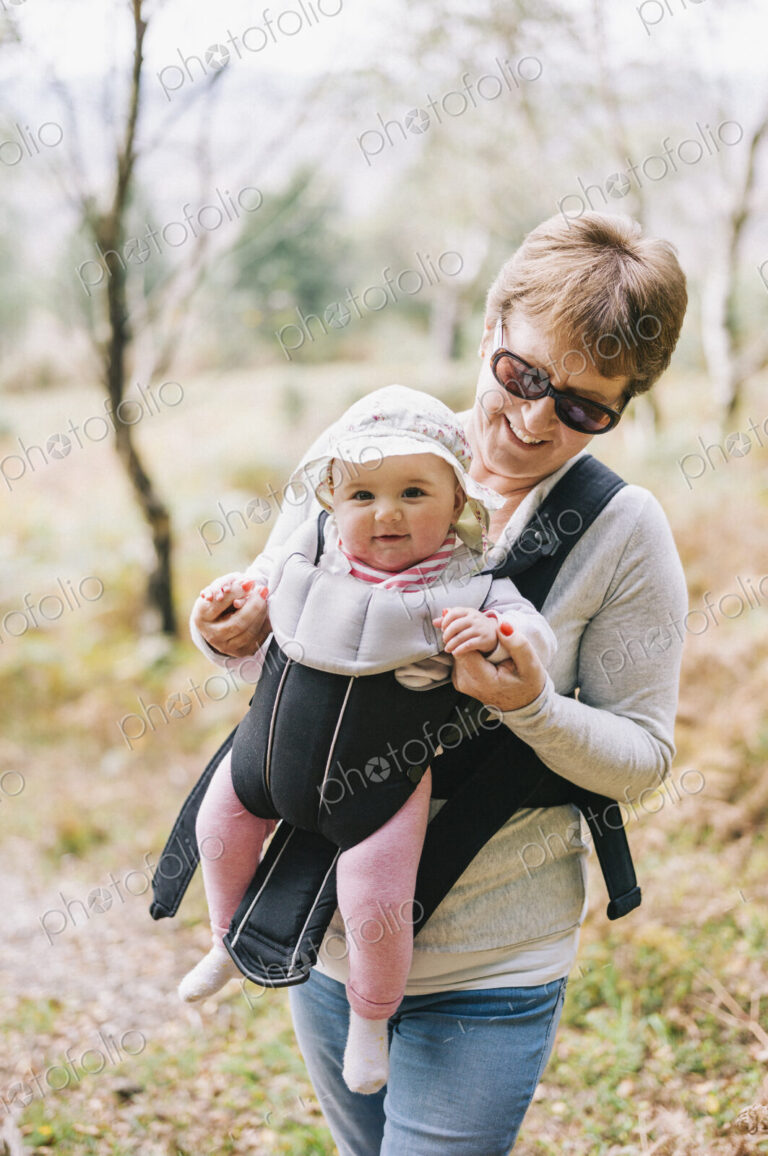 A woman wearing sunglasses carrying a baby wearing a white sunhat in a front baby sling.