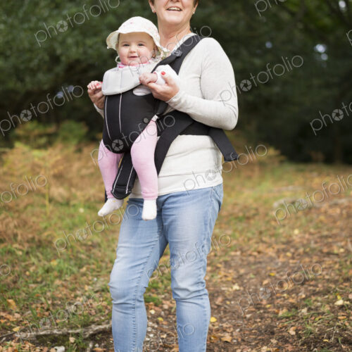 A woman wearing sunglasses carrying a baby wearing a white sunhat in a front baby sling.