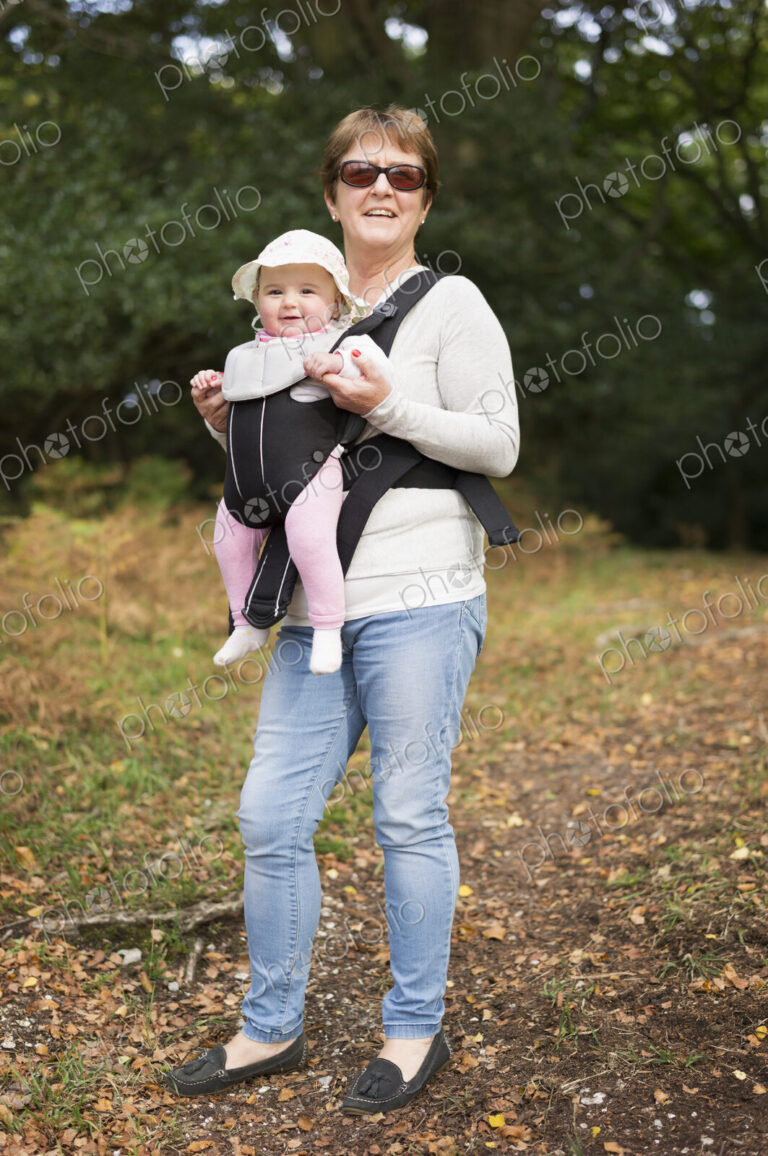 A woman wearing sunglasses carrying a baby wearing a white sunhat in a front baby sling.