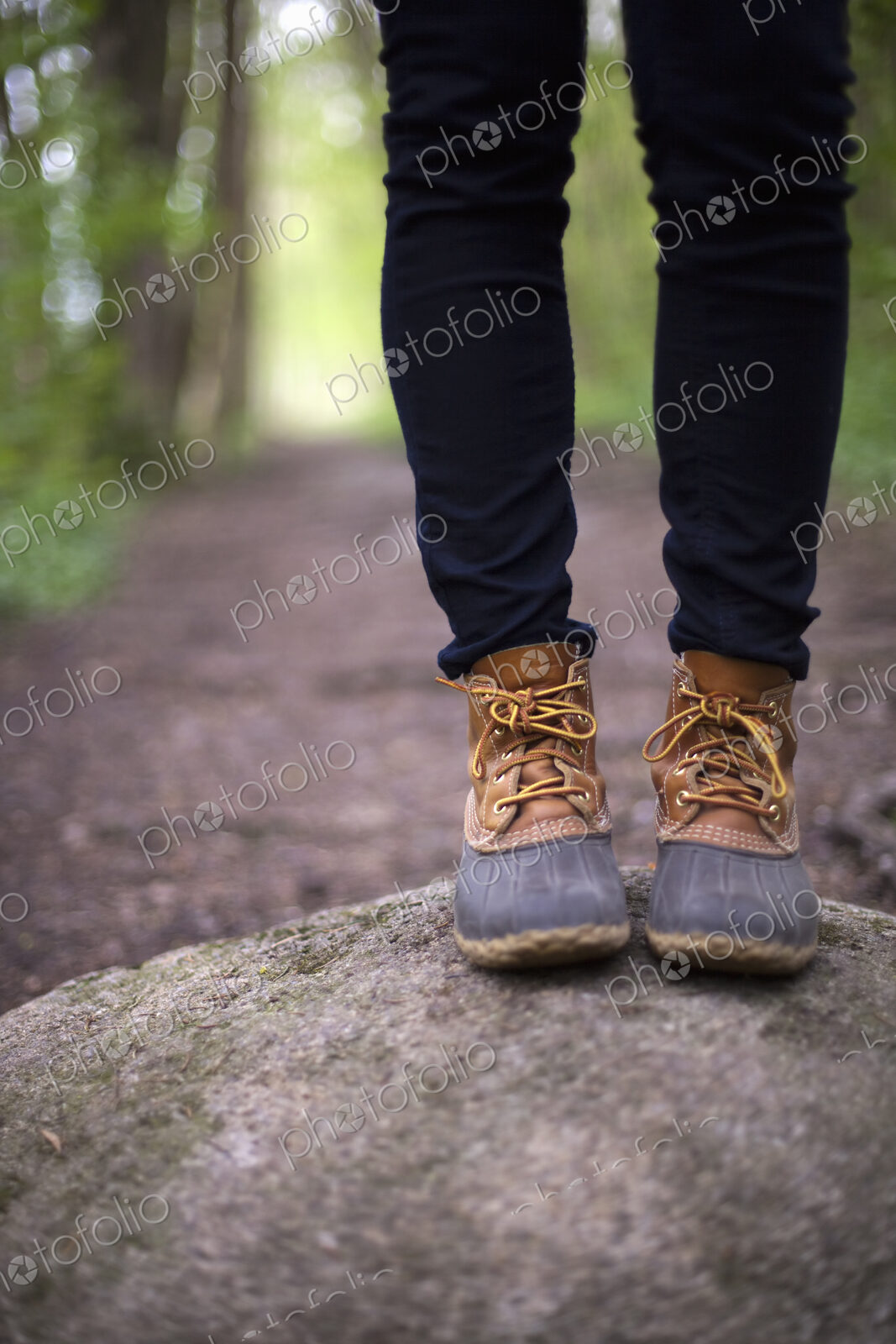 A woman wearing walking boots and jeans, standing on a stone beside a path in woodland.
