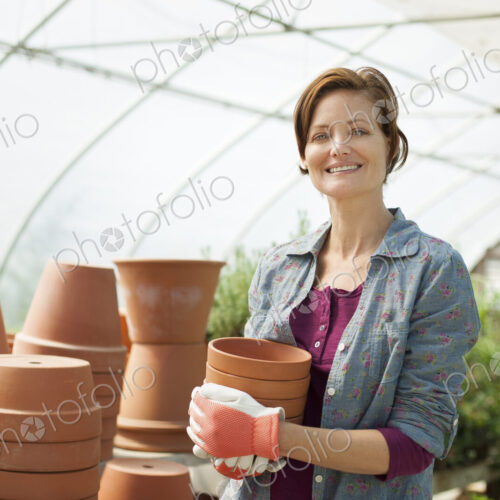 A woman wearing workgloves carrying terracotta plant pots in a large greenhouse on an… A woman wearing workgloves carrying terracotta plant pots in a large greenhouse on an…
