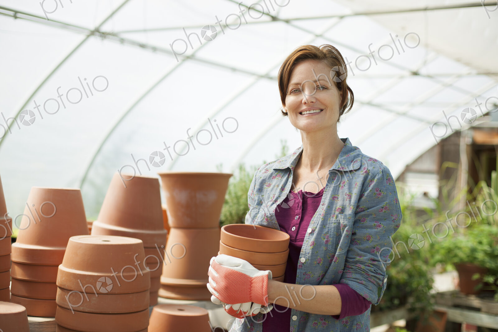 A woman wearing workgloves carrying terracotta plant pots in a large greenhouse on an…