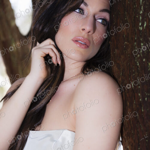 A woman with bare shoulders leaning by a tree outdoors.
