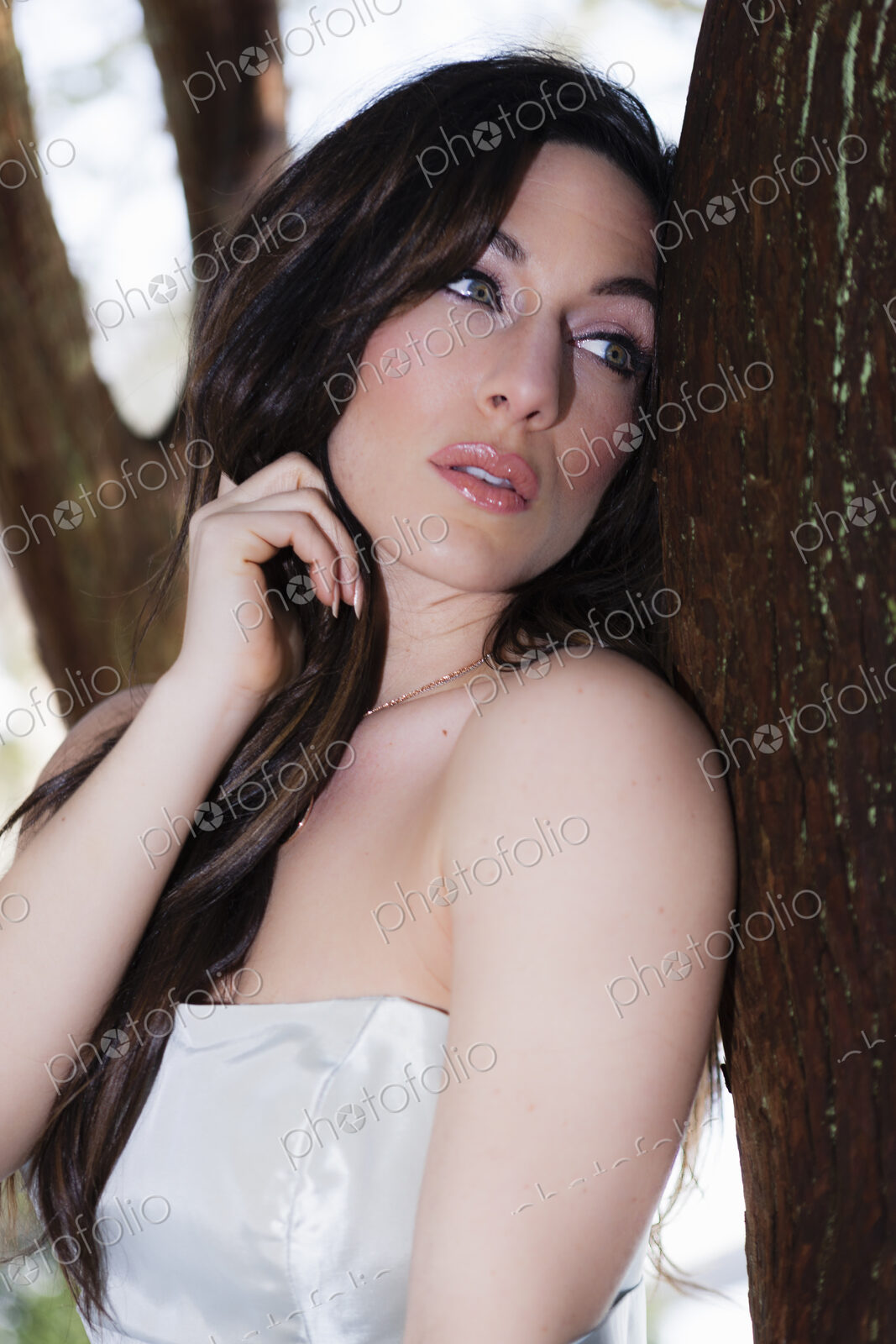 A woman with bare shoulders leaning by a tree outdoors.