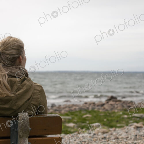 A woman with long blonde hair wearing a brown hooded coat, seated on a bench looking… A woman with long blonde hair wearing a brown hooded coat, seated on a bench looking…
