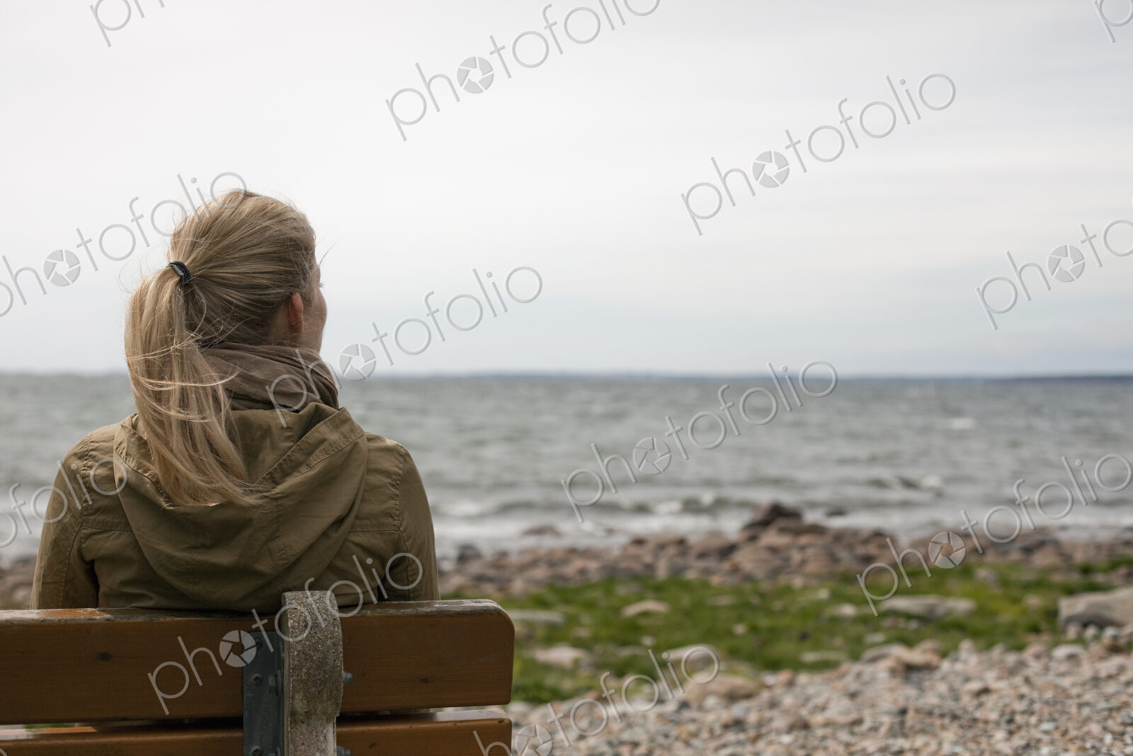A woman with long blonde hair wearing a brown hooded coat, seated on a bench looking…