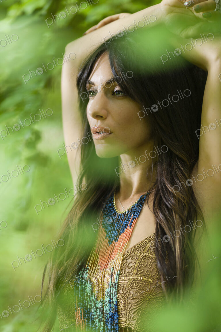 A woman with long brown hair outdoors in woodland with her arms above her head.